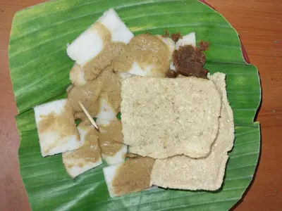 Porsi Cabuk Rambak Solo berisi irisan ketupat beras dengan saus wijen pucat dan kerupuk karak beras di atas alas daun pisang.