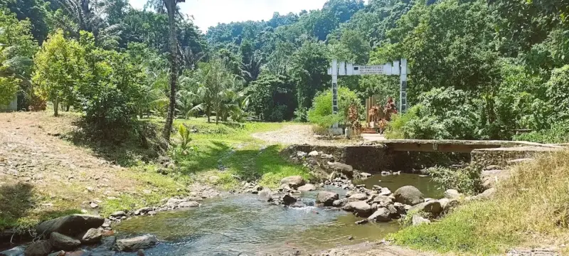 Gerbang masuk wisata Air Terjun Tunan Talawaan dengan jembatan kecil yang melintasi aliran sungai berbatu dangkal, dikelilingi pepohonan kelapa dan vegetasi hijau yang asri.
