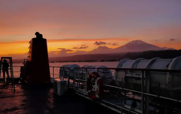 Itinerary Jakarta Bali via Kereta Api. Kapal ferry ASDP bersandar di Pelabuhan Ketapang Banyuwangi di bawah terik matahari pesisir, siap menyeberangkan penumpang menuju Gilimanuk Bali.
